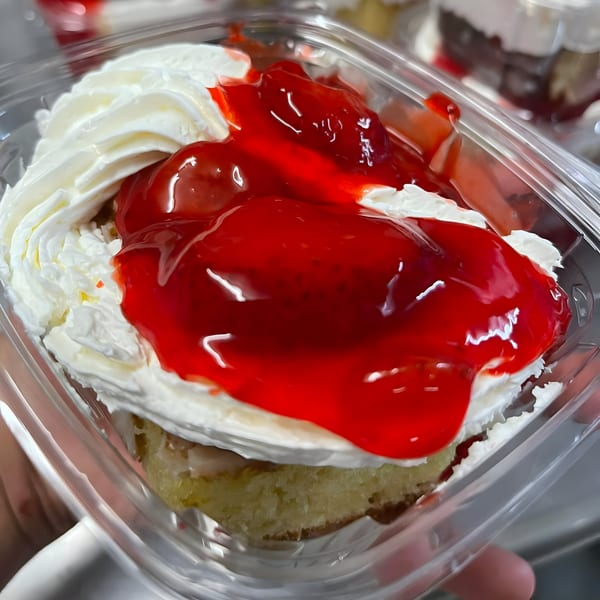 A clear plastic container holds a dessert with white whipped cream and bright red strawberry topping.